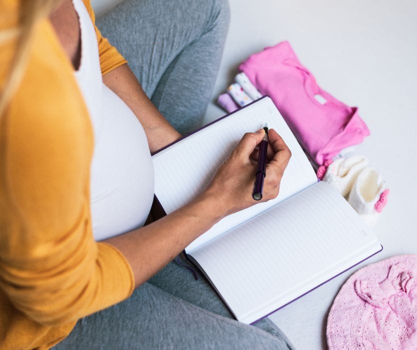 Pregnant woman sitting on the floor writing in a notebook next to pink baby clothes and booties.