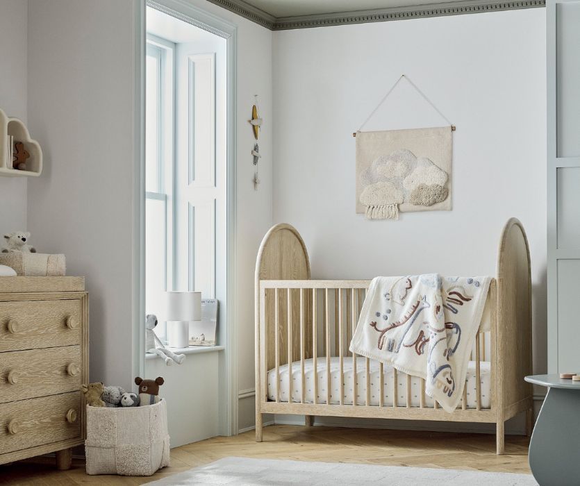 Modern baby nursery with a light wood crib, animal-themed blanket, matching dresser, and cloud wall hanging.