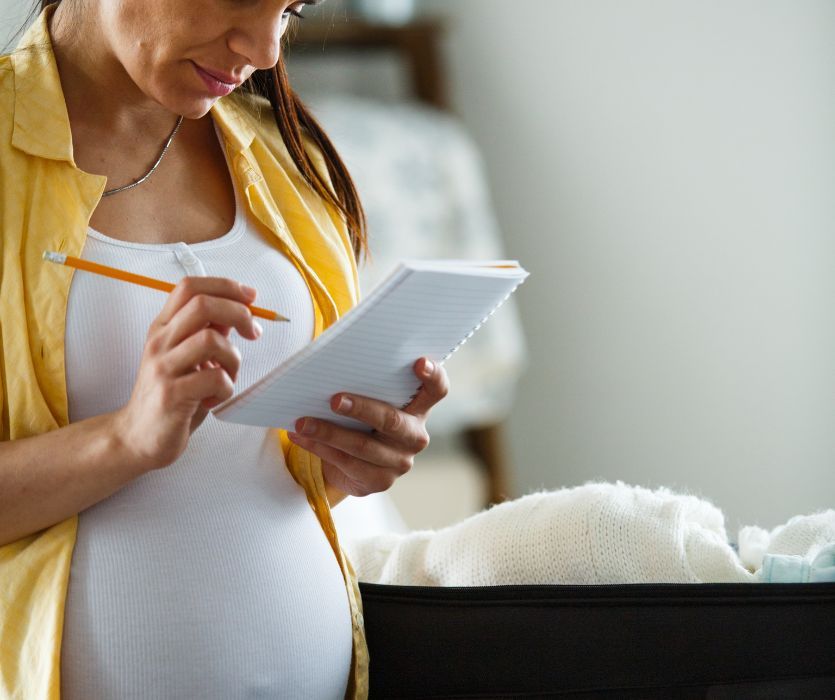 Pregnant woman in a yellow shirt writing on a notepad while packing a suitcase with baby clothes.