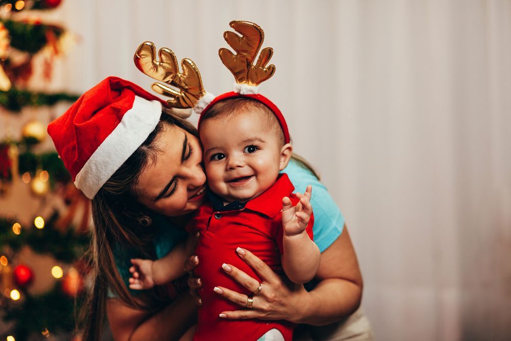 Smiling baby wearing reindeer antlers being held by a woman in a Santa hat next to a Christmas tree.