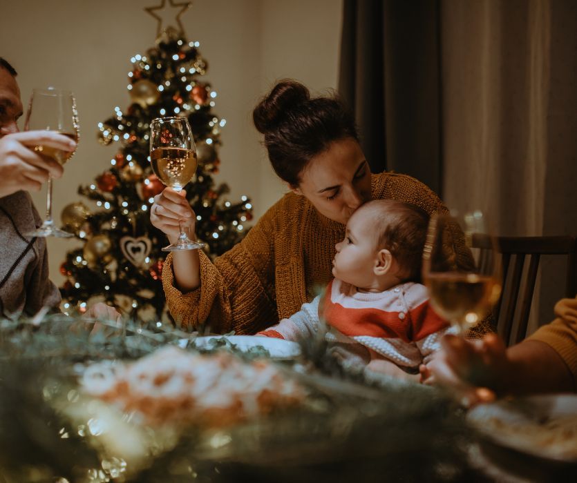 A woman kisses a baby's head while holding a wine glass at a Christmas dinner table near a lit tree.