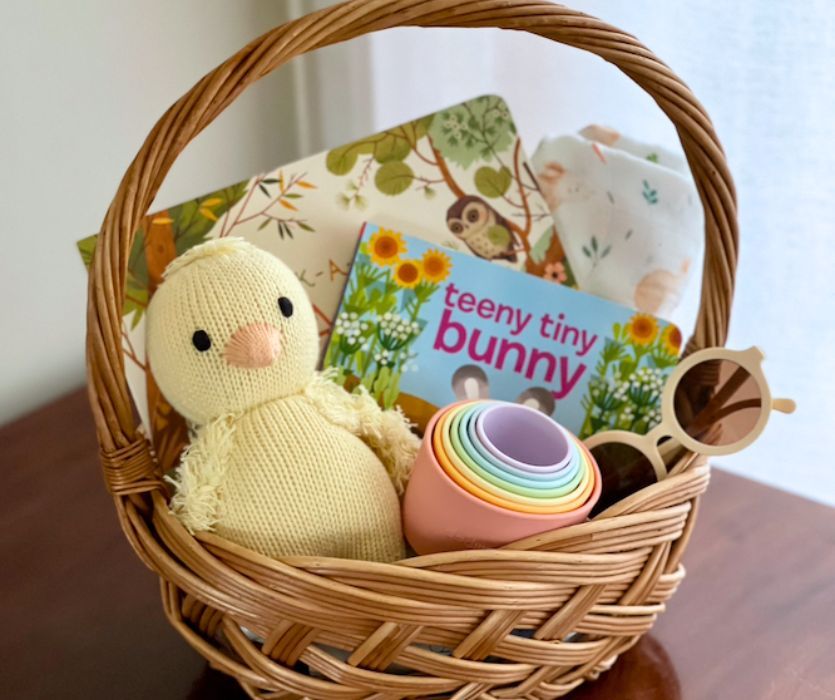 Baby Easter basket with a knit chick, board books, stacking cups, sunglasses, and a swaddle blanket.