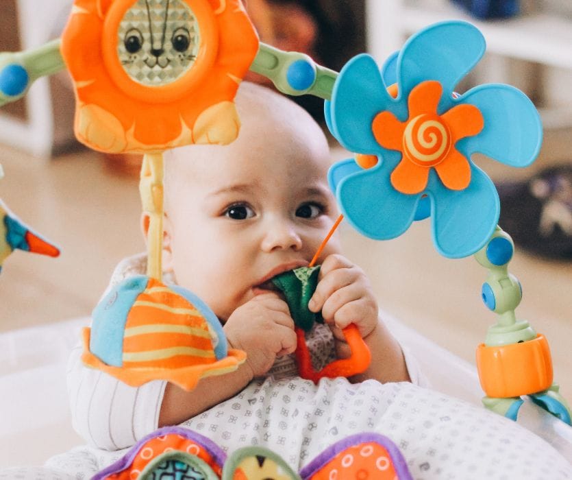 A baby chewing on a colorful hanging toy attached to a plastic activity arch.