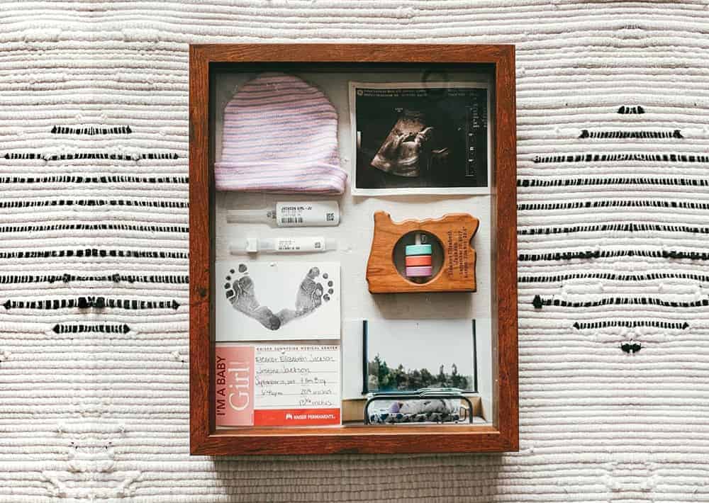 Baby keepsakes in a wooden shadow box including a striped hat, ultrasound, footprints, hospital bands, and a birth card.