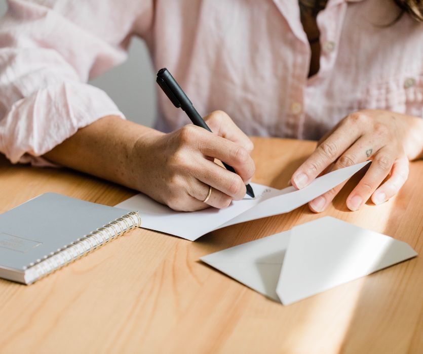 A person in a pink shirt writes a message inside a white greeting card with a black pen on a wooden desk.