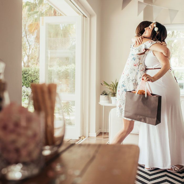 A woman in a white dress hugging a guest who is holding a gift bag at a baby shower.