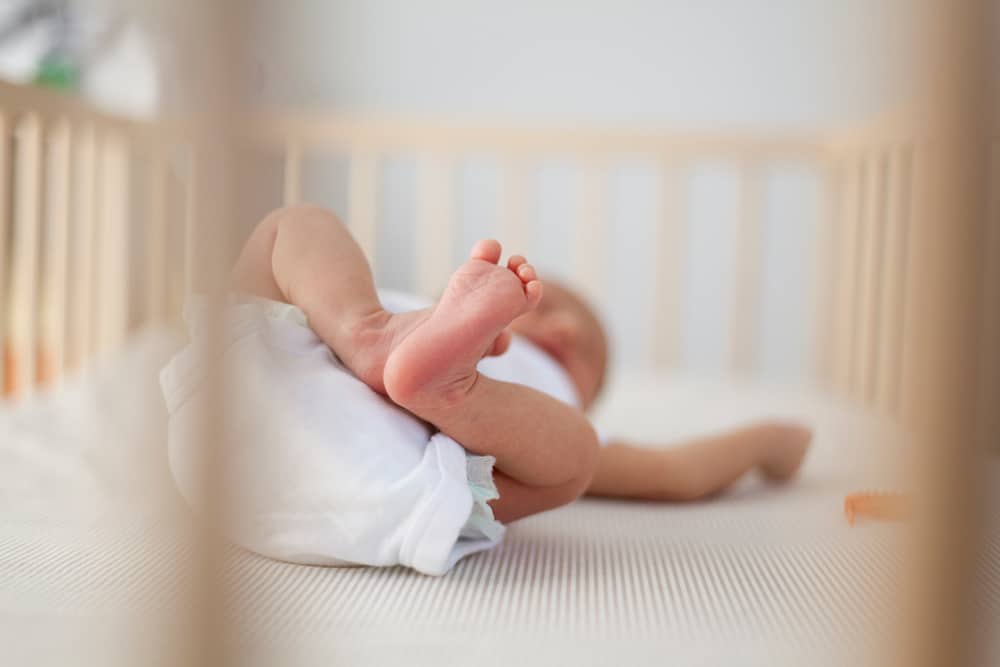 Newborn baby lying on back in a wooden crib with a firm, flat mattress.