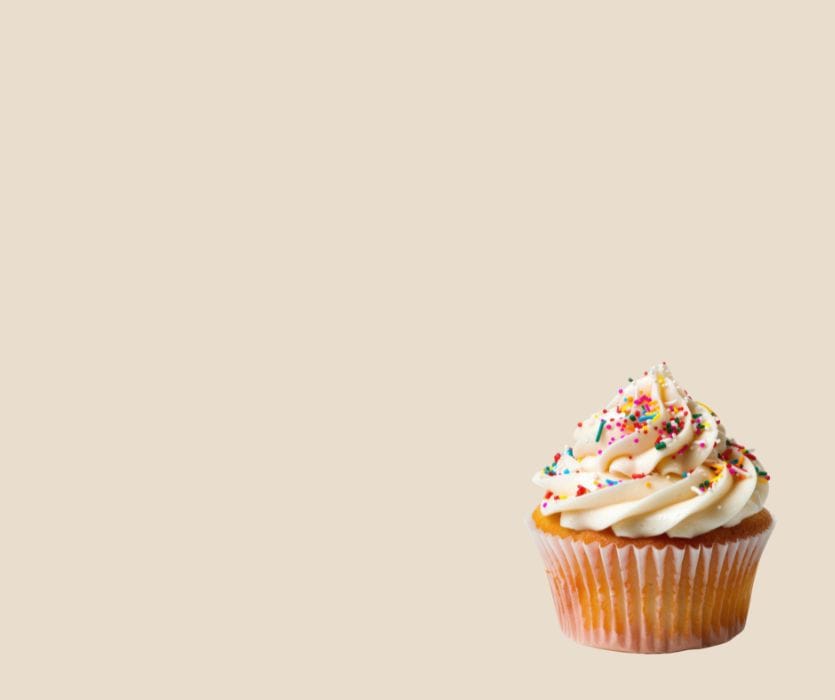 Vanilla cupcake with white frosting and multi-colored rainbow sprinkles against a light tan background.