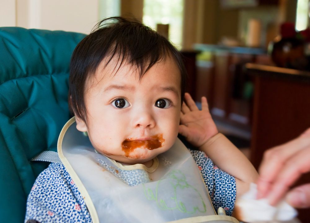 A baby sitting in a high chair with brown puree food smeared around their mouth and on their bib.