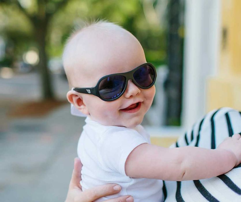 A smiling baby wearing black sunglasses and a white t-shirt held by an adult outdoors.