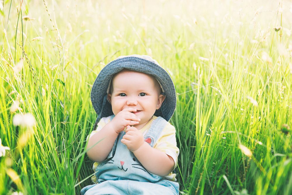 Smiling baby wearing a denim sun hat and overalls sitting in a field of tall green grass.
