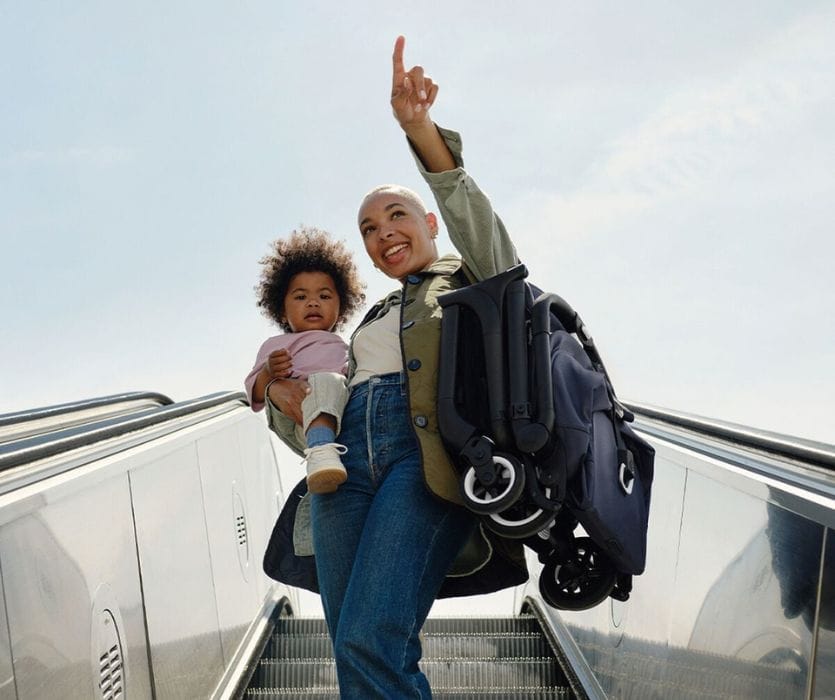 A woman holding a toddler and a folded compact stroller while standing on an escalator.