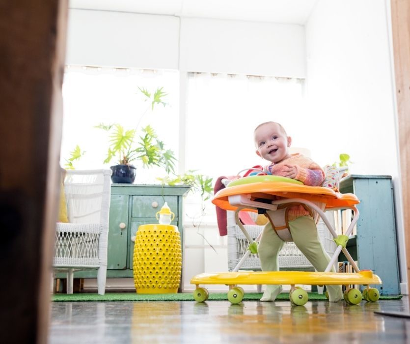 Smiling baby in a vibrant orange and yellow baby walker in a sunlit room with plants and colorful furniture.