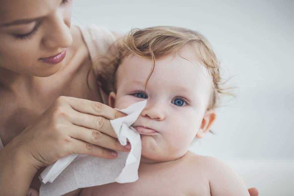 A woman uses a white baby wipe to clean the nose and face of a blue-eyed infant.