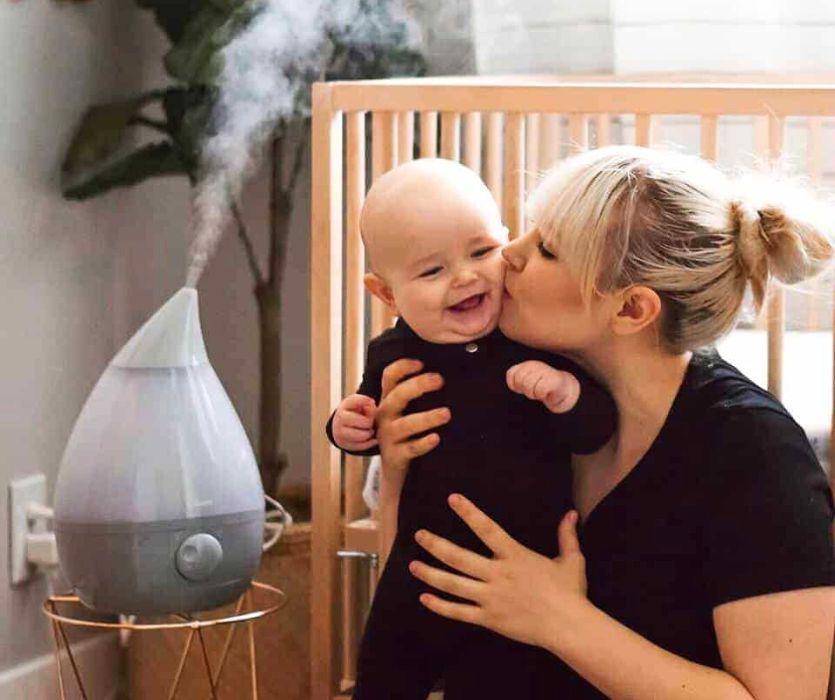 A woman kisses a smiling baby's cheek next to a teardrop-shaped humidifier emitting mist in a nursery.