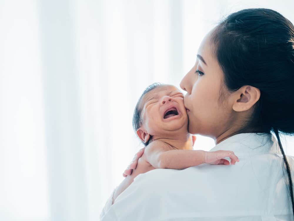 A woman kisses the cheek of a crying newborn baby held against her shoulder.