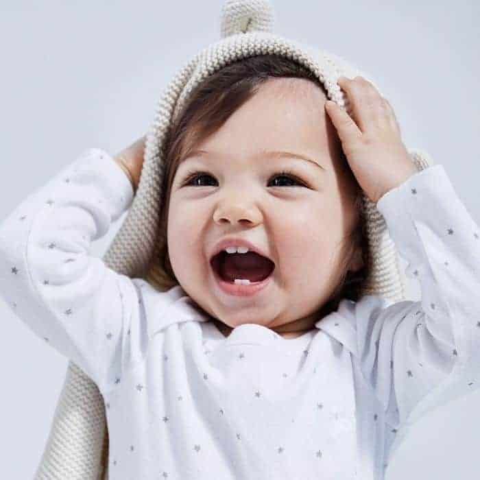 Smiling baby in a star-patterned shirt holding a garter stitch knit hat over their head.
