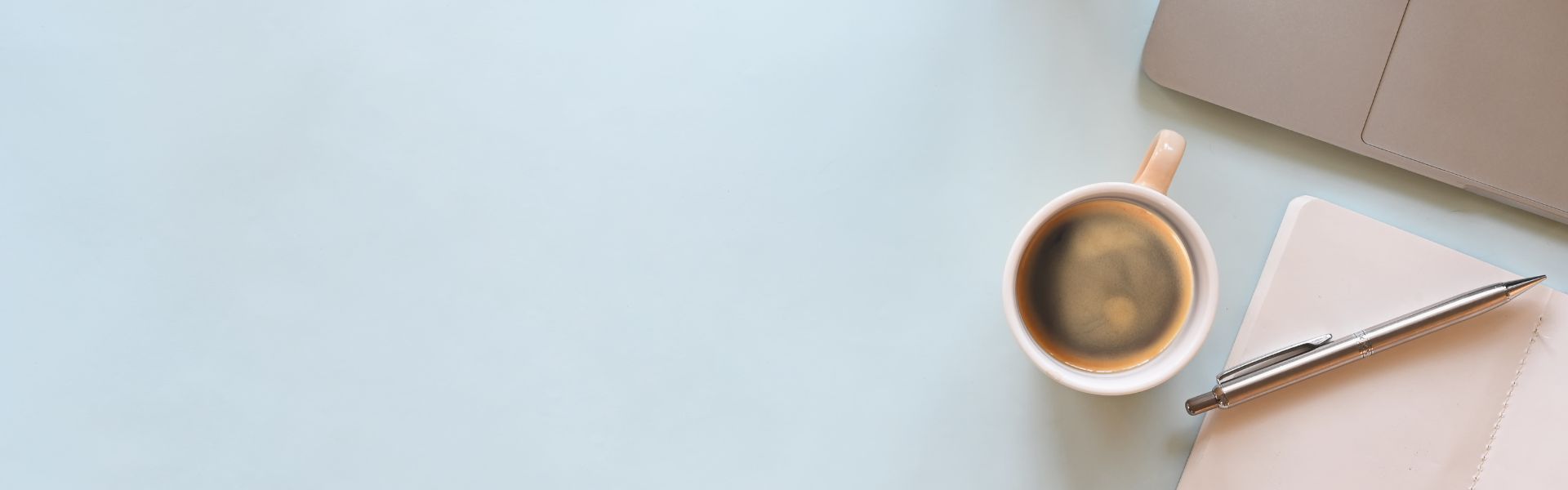Top-down view of a coffee cup, silver pen, notebook, and laptop on a light blue desk with copy space.