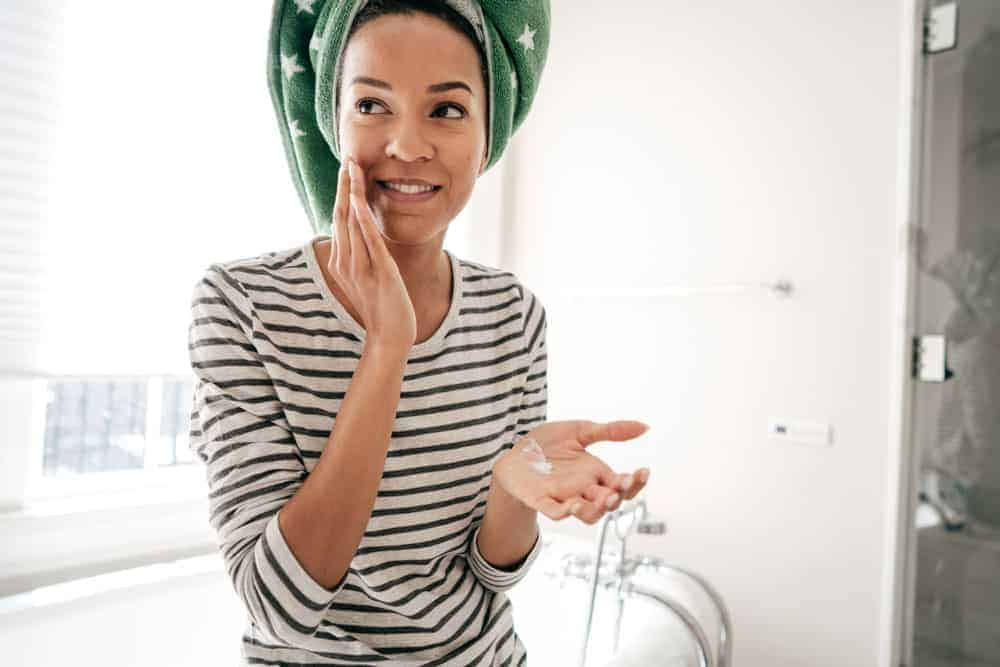 Woman with a green hair towel applying a facial cream to her cheek in a bathroom.