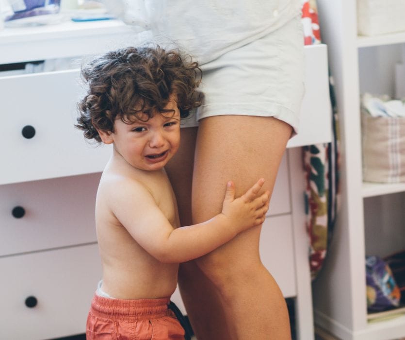 A curly-haired toddler crying and clinging to a parent's leg in a bedroom.