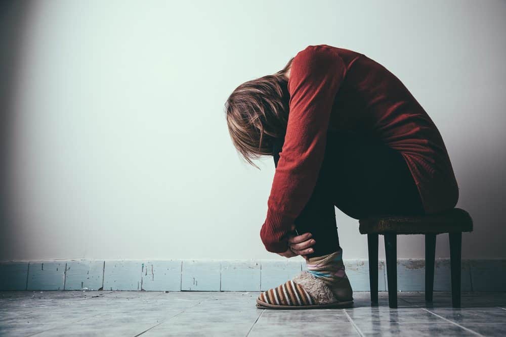 A person in a red sweater sits hunched over on a stool, head buried in their arms, representing grief and mourning.