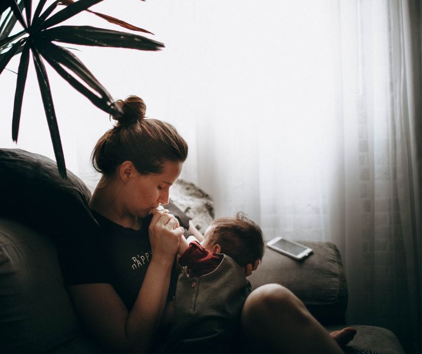A woman sitting on a couch, holding and kissing the hand of an infant in her lap.
