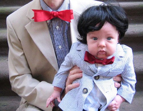 Hand holding a baby dressed in a seersucker suit, red bowtie, and black wig as a ventriloquist dummy.