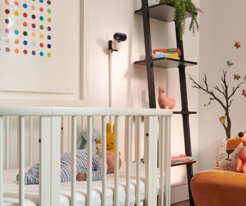 A wall-mounted baby monitor camera overlooks a sleeping baby in a white crib inside a decorated nursery.
