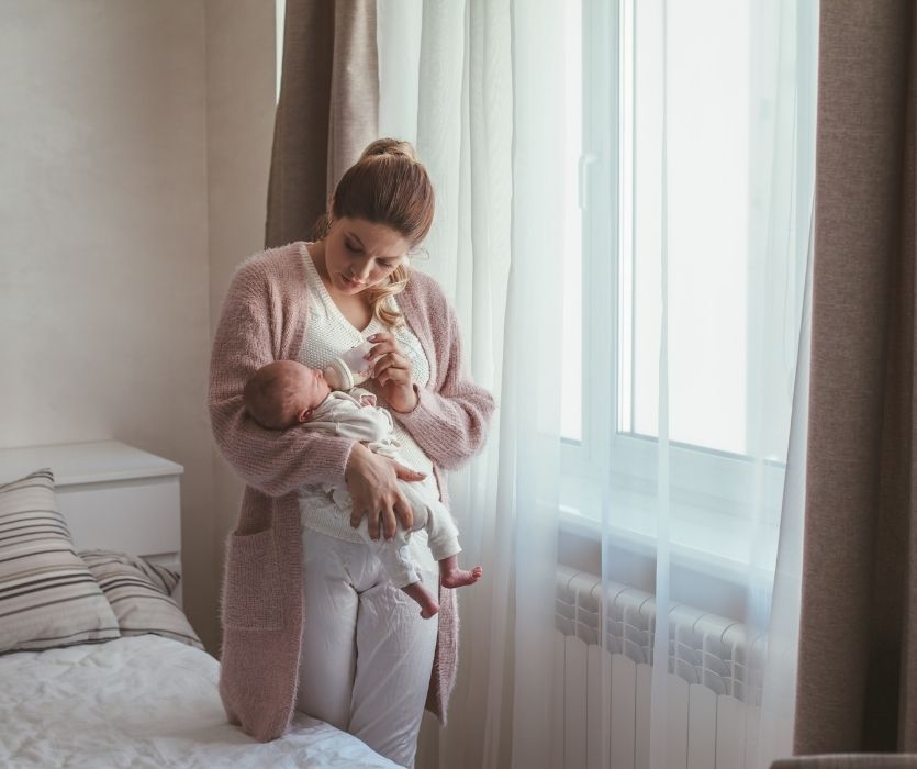 A mother in a pink cardigan bottle-feeding her newborn baby while standing by a bedroom window.