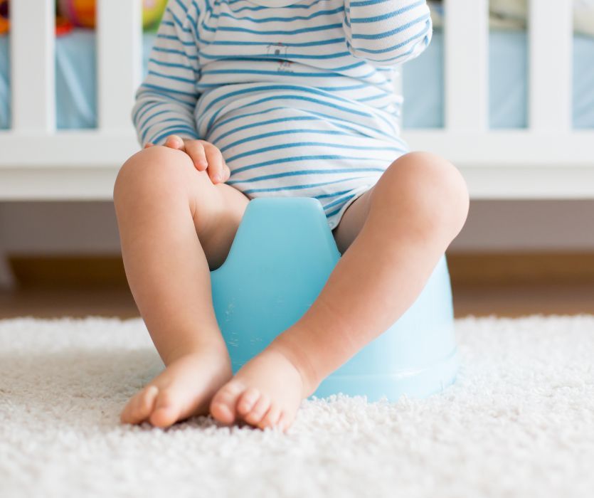 A toddler in a striped onesie sits on a small blue plastic potty chair on a white rug.