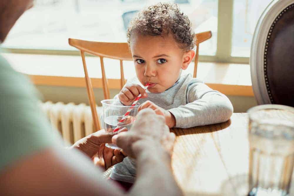 An adult helps a young child drink water through a striped straw from a clear plastic cup at a wooden table.