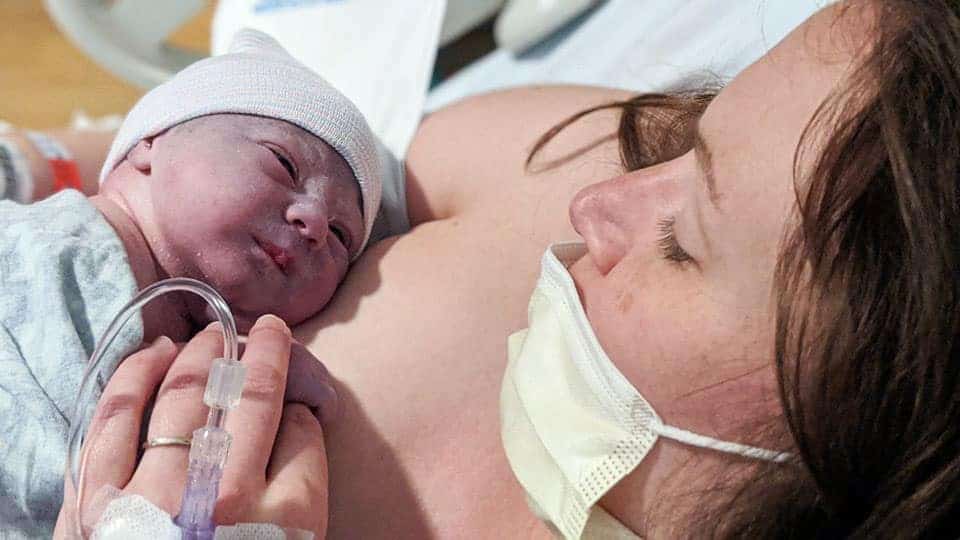 Close-up of a woman in a medical mask holding a newborn baby against her chest in a hospital setting.