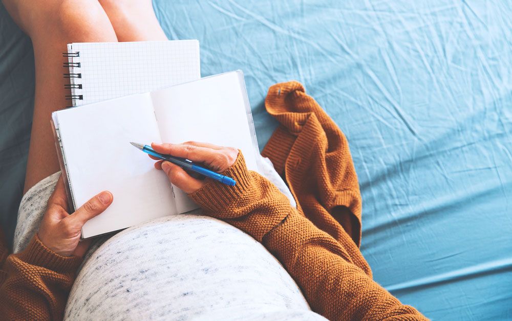 Top-down view of a pregnant person sitting on a bed and writing in a blank notebook with a blue pen.