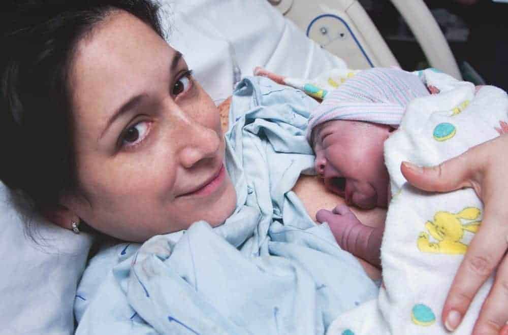 A mother in a hospital bed smiling while holding her crying newborn baby wrapped in a blanket.