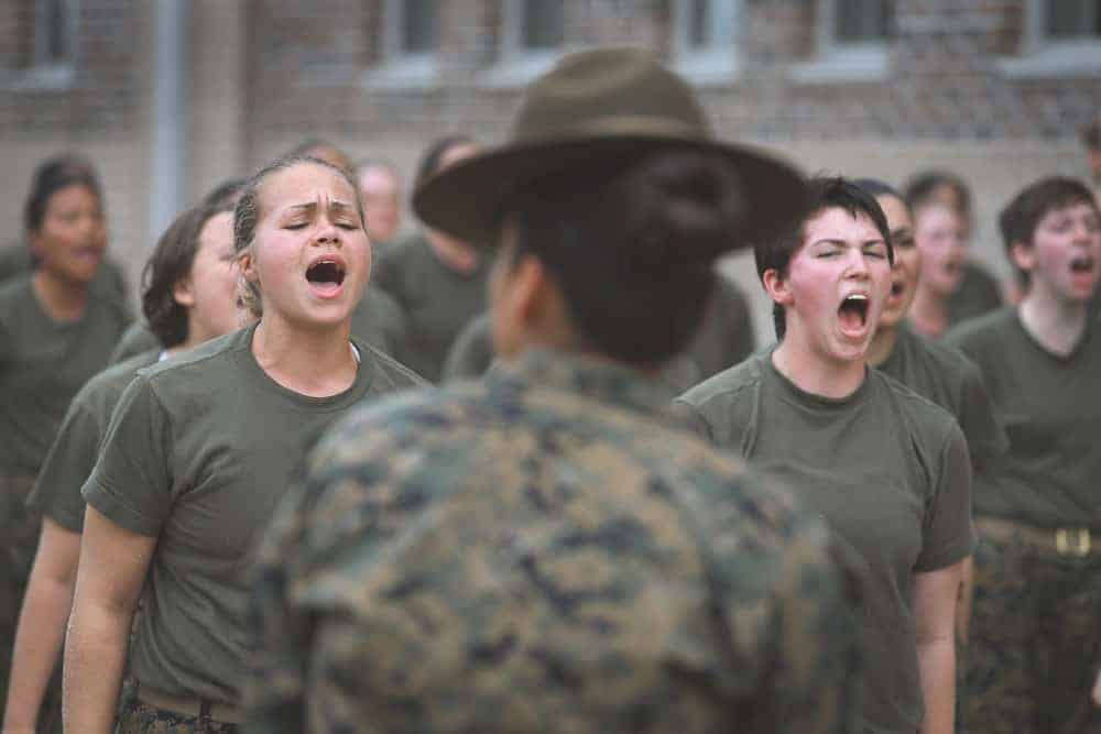 Female recruits in olive shirts shout during training as a drill instructor in a campaign hat stands with her back to camera.
