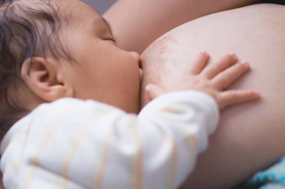 Close-up of a baby latched onto a large breast during breastfeeding, with the baby's hand resting on the breast.