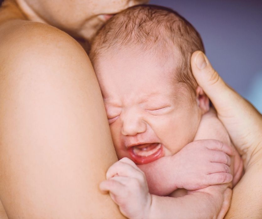 A crying newborn baby being held close against a person's chest.