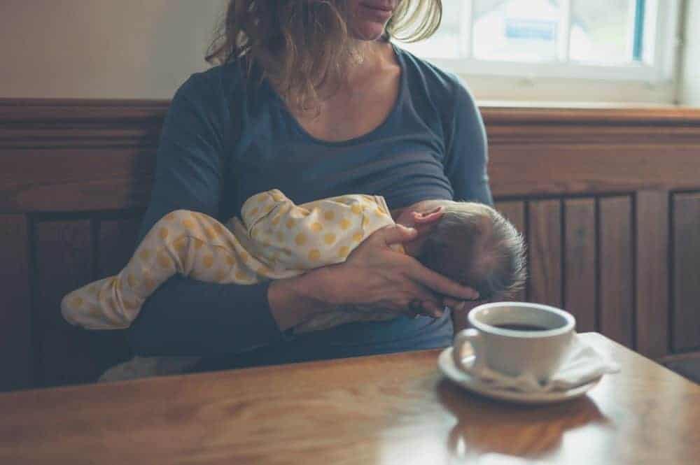 A woman breastfeeds her infant while sitting at a table with a cup of coffee.