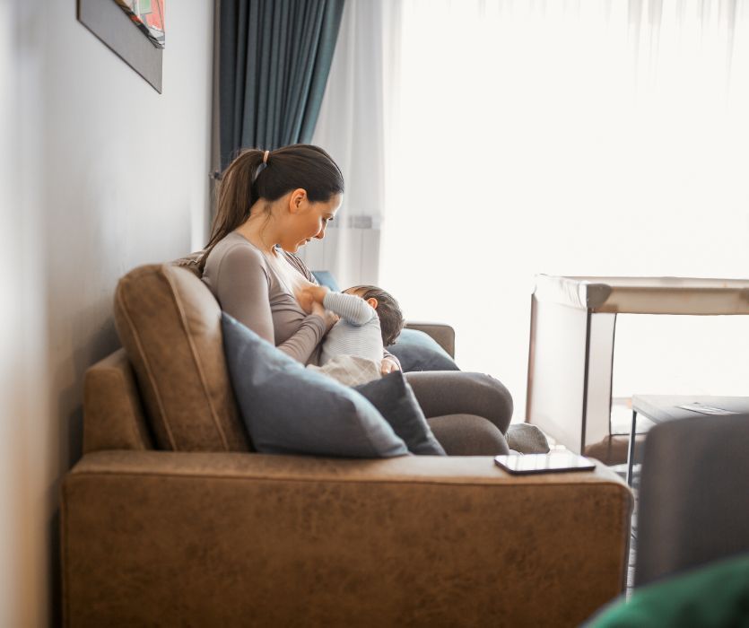 A woman sitting on a brown couch while breastfeeding her baby in a brightly lit living room.