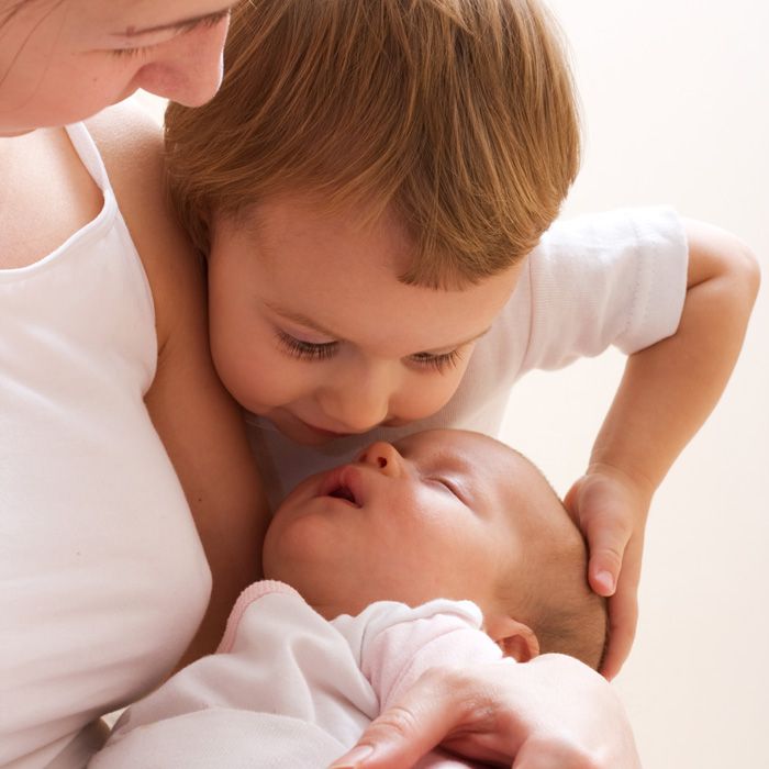 A mother and young child leaning over to look at a sleeping newborn baby.