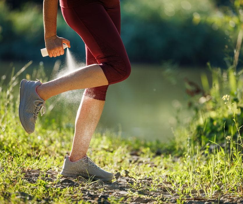 A person in athletic wear applying bug repellent spray to their legs while standing outdoors in a grassy area.