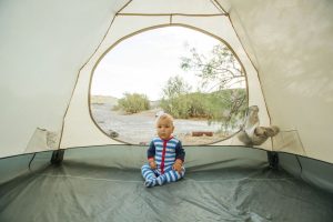 A baby wearing blue and white striped pajamas sits inside a tent looking out at a desert campsite.