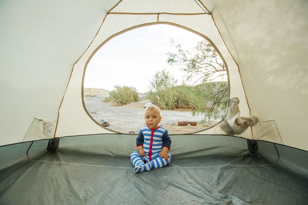 A baby wearing blue and white striped pajamas sits inside a tent looking out at a desert campsite.