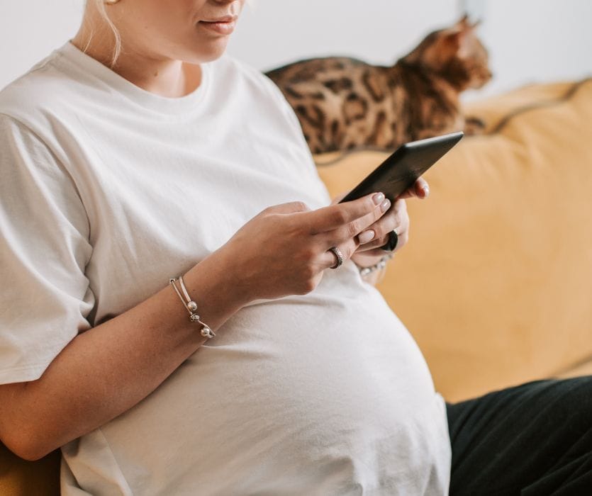 A pregnant woman sitting on a couch and looking at a tablet, with a Bengal cat in the background.