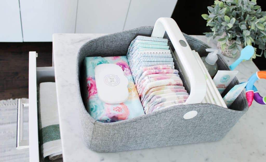 Grey fabric diaper caddy organized with floral wipes, patterned diapers, lotion, and baby supplies on a white counter.