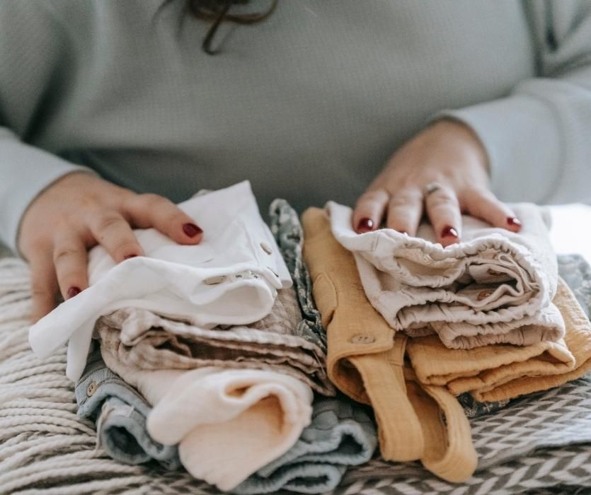 A person’s hands resting on stacks of folded baby clothes including onesies and trousers.