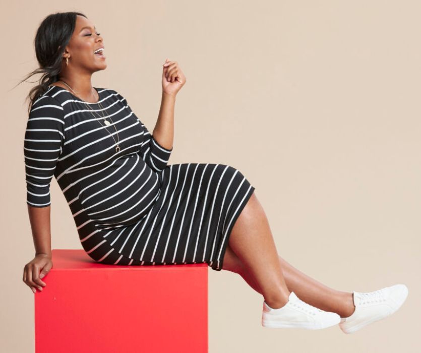 Pregnant woman laughing in a black and white striped bodycon maternity dress and white sneakers.