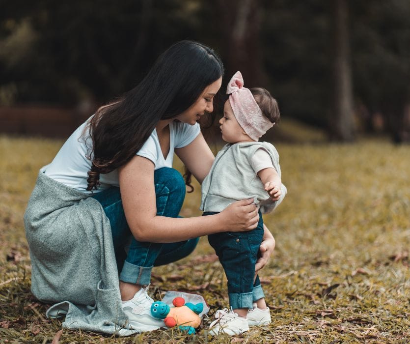 A woman crouches in a field and smiles while holding a standing baby wearing a pink bow headband.