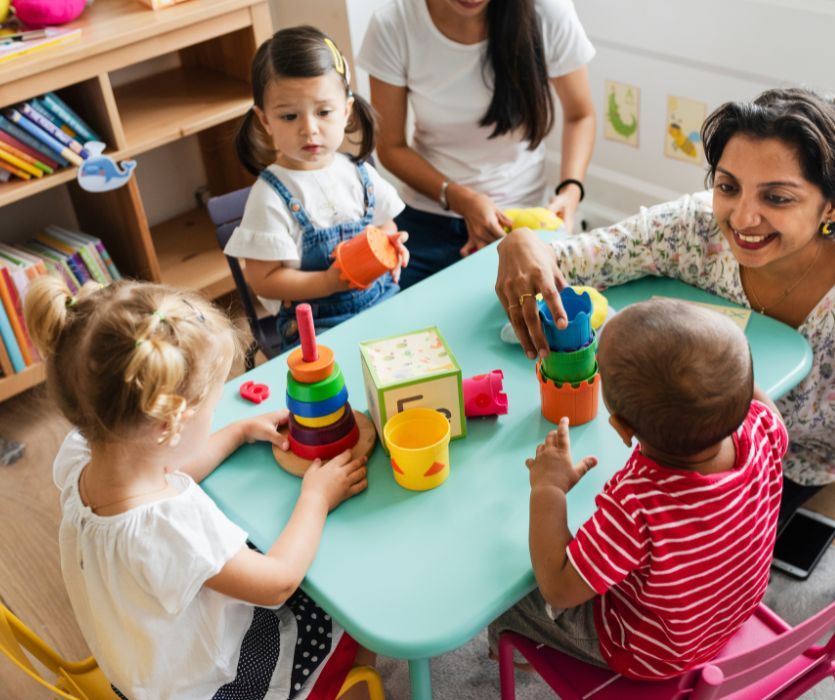 Two teachers and four young children playing with colorful blocks and stacking toys at a blue table in a daycare.