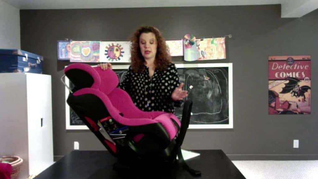 A woman stands behind a pink and black Clek Fllo convertible car seat placed on a black table.